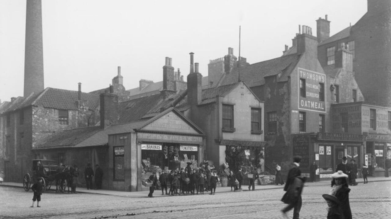 Black and white photo of a busy street corner with shops, people, horse-drawn cart, and tall brick chimneys in an industrial-era city.