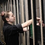 A person in black clothing adjusts stage rigging ropes backstage in a theater, surrounded by multiple vertical ropes and weights.