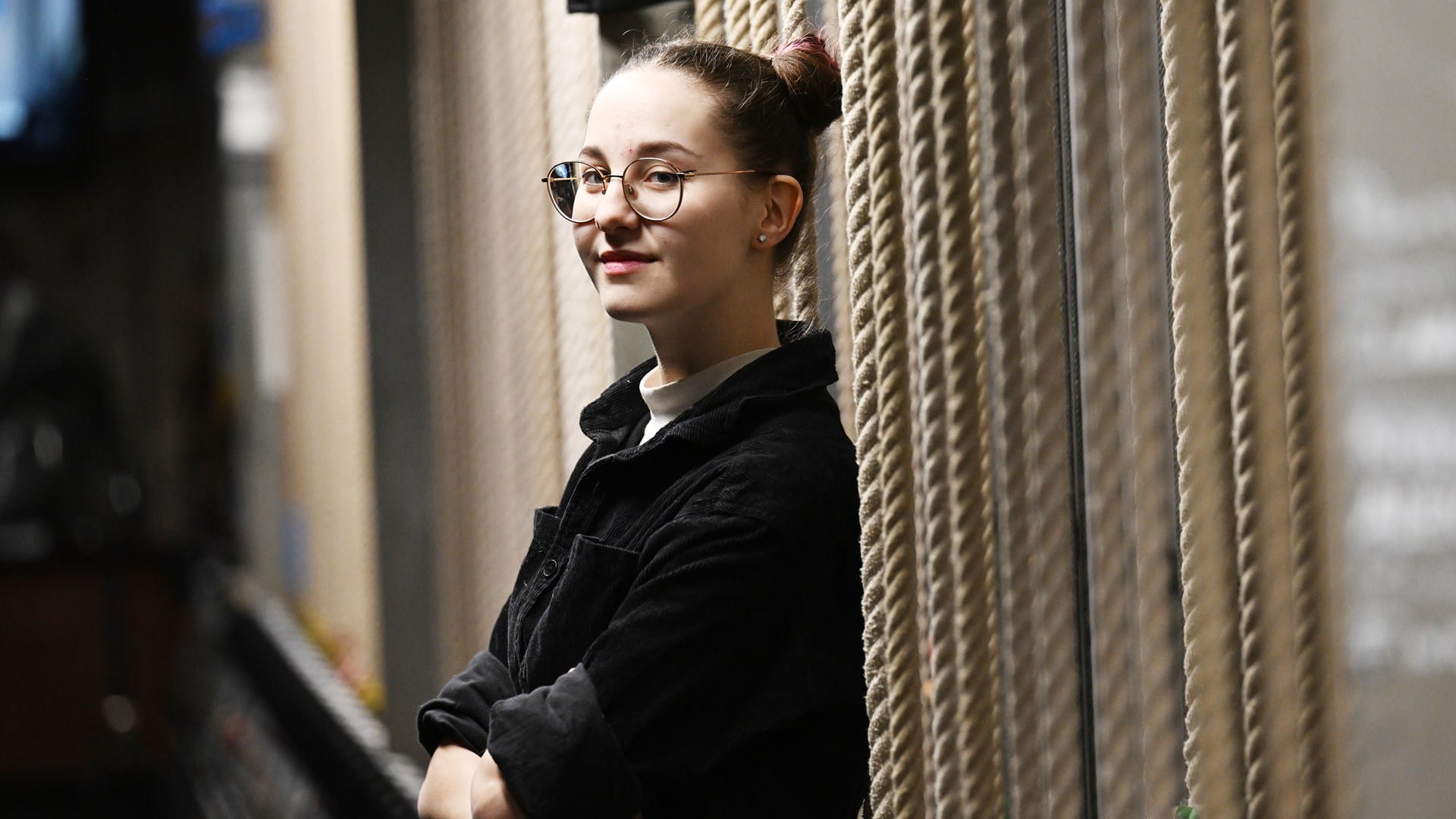 A person with glasses and hair in a bun leans against a wall with thick ropes, arms crossed, looking at the camera.