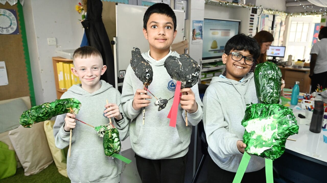 Three boys in grey hoodies stand indoors, each holding handmade animal models crafted from painted materials and sticks, smiling at the camera in a classroom setting.