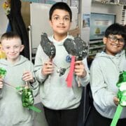 Three boys in grey hoodies stand indoors, each holding handmade animal models crafted from painted materials and sticks, smiling at the camera in a classroom setting.