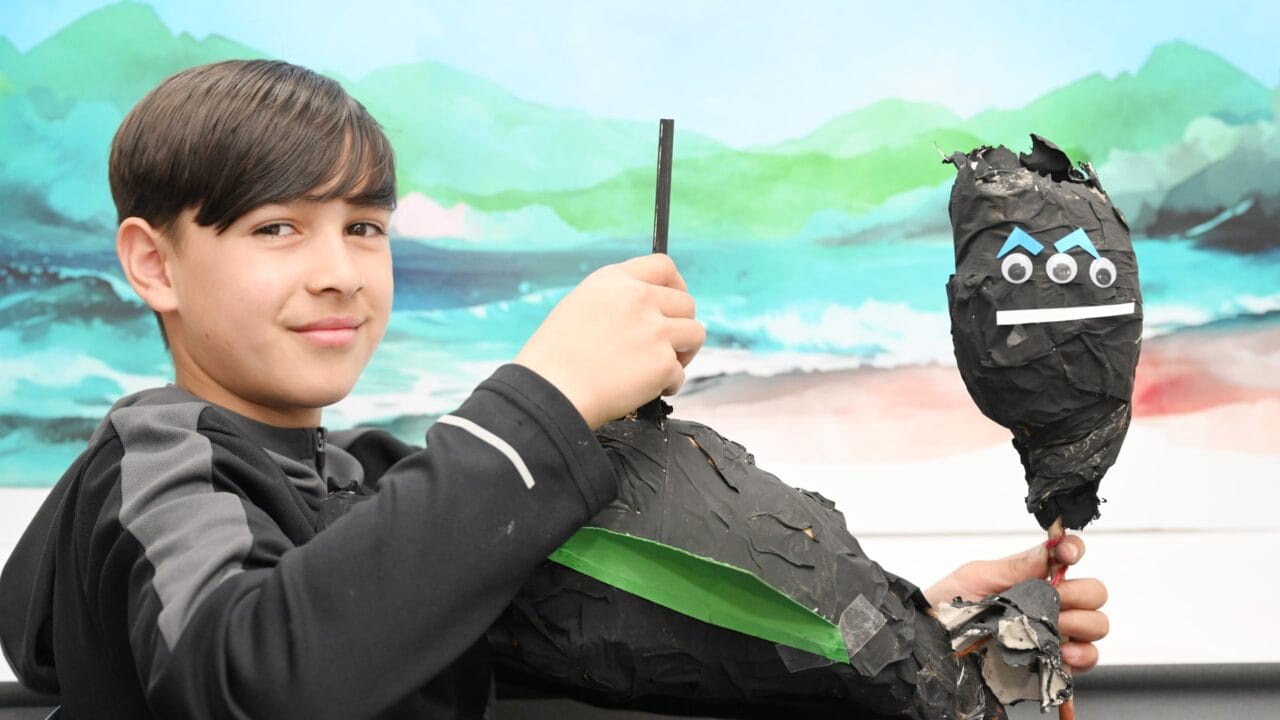 A boy holds a homemade black papier-mâché puppet with googly eyes and blue paper eyebrows, sitting in front of a painted landscape background.
