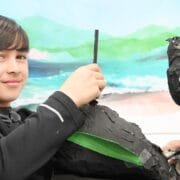A boy holds a homemade black papier-mâché puppet with googly eyes and blue paper eyebrows, sitting in front of a painted landscape background.