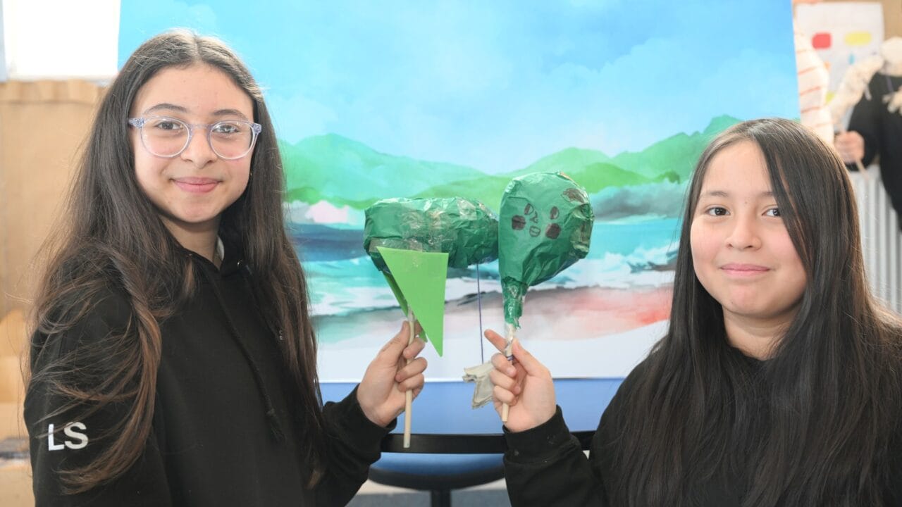 Two girls stand in front of a painted landscape backdrop, each holding a green, heart-shaped craft on a stick and smiling at the camera.