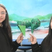 Two girls stand in front of a painted landscape backdrop, each holding a green, heart-shaped craft on a stick and smiling at the camera.