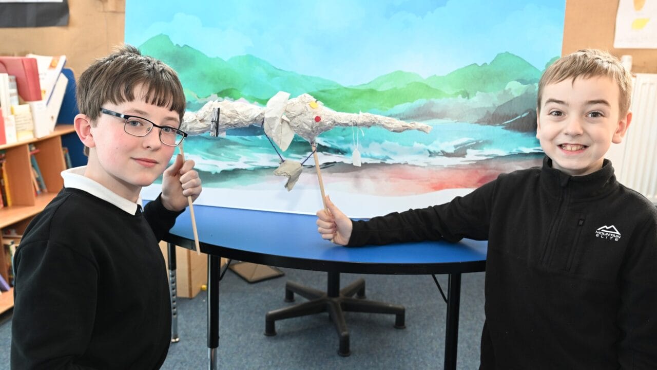 Two boys stand by a table, holding sticks to control a paper puppet in front of a painted backdrop of mountains and a river.