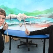 Two boys stand by a table, holding sticks to control a paper puppet in front of a painted backdrop of mountains and a river.