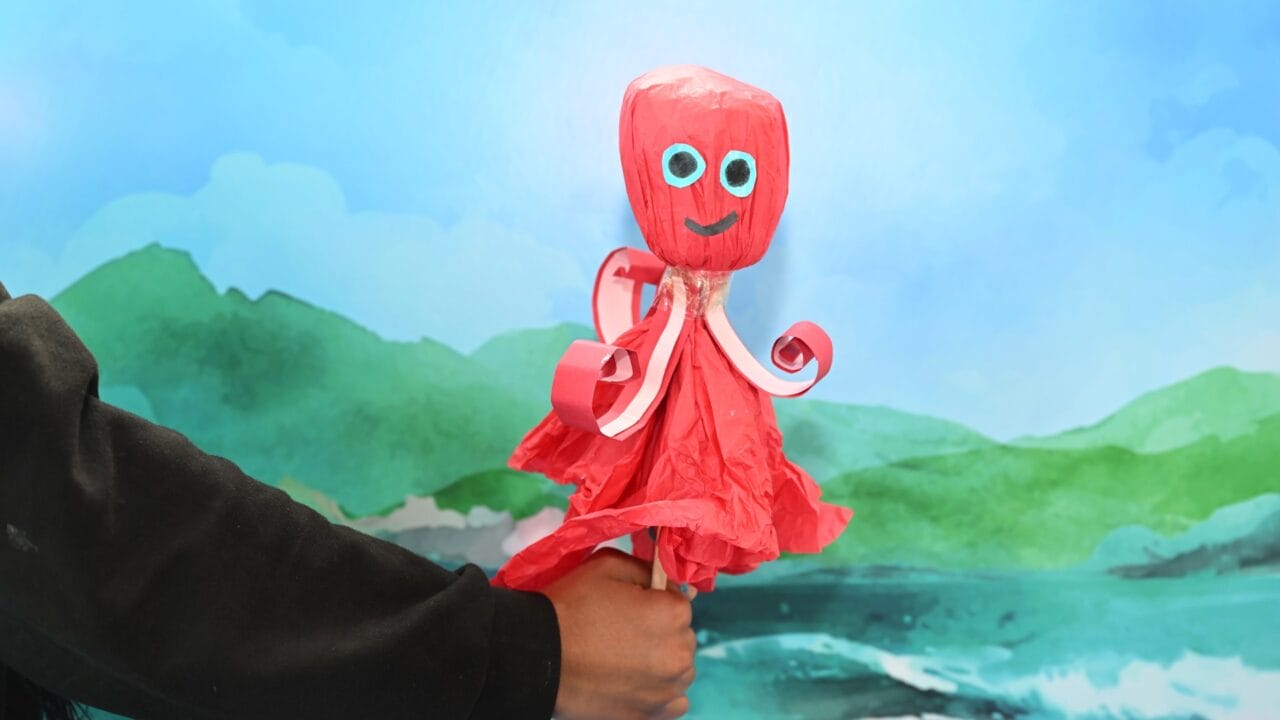 A hand holds a red octopus craft made from paper materials, posed in front of a painted backdrop of mountains and water.