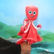A hand holds a red octopus craft made from paper materials, posed in front of a painted backdrop of mountains and water.