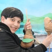 A smiling child in a black coat holds up two paper mache puppets in front of a painted backdrop of mountains and water.