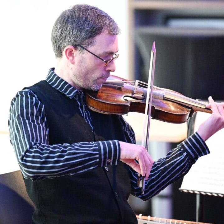 A man wearing glasses plays a violin while seated indoors, with sheet music on a stand in front of him.