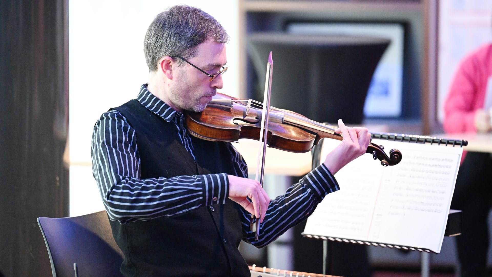 A man wearing glasses plays a violin while seated indoors, with sheet music on a stand in front of him.