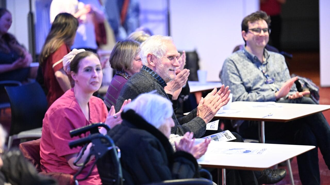 People of various ages, including a woman in scrubs and an elderly person in a wheelchair, sit at tables and applaud during an indoor event.