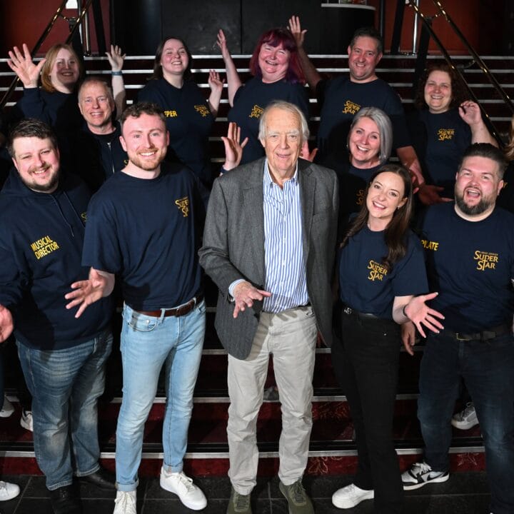 A group of people, most wearing matching “Jesus Christ Superstar” shirts, pose together on a staircase with an older man in the centre. All are smiling and gesturing with open hands.