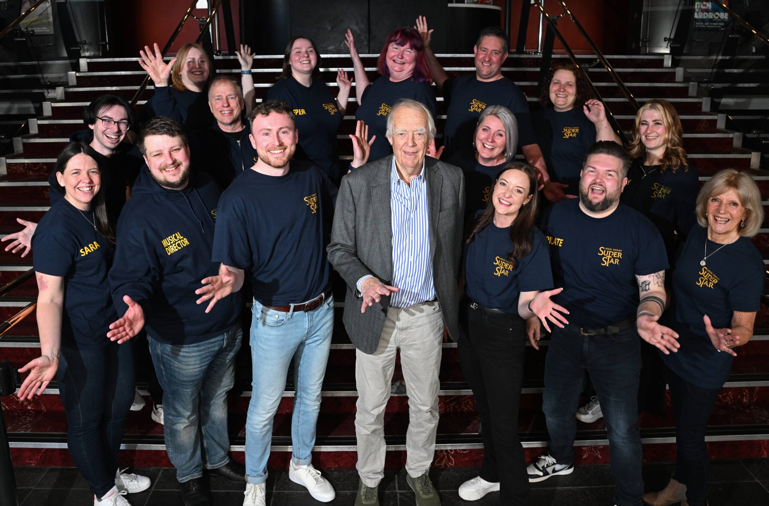 A group of people, most wearing matching “Jesus Christ Superstar” shirts, pose together on a staircase with an older man in the centre. All are smiling and gesturing with open hands.
