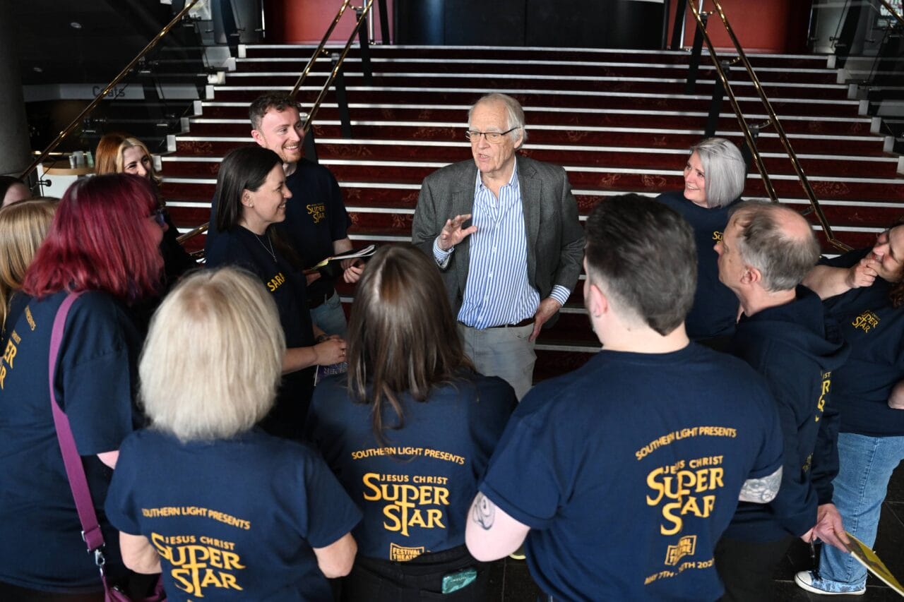 An older man speaks to a group of people wearing "Jesus Christ Superstar" shirts in front of a staircase.