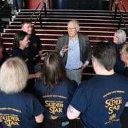 An older man speaks to a group of people wearing "Jesus Christ Superstar" shirts in front of a staircase.