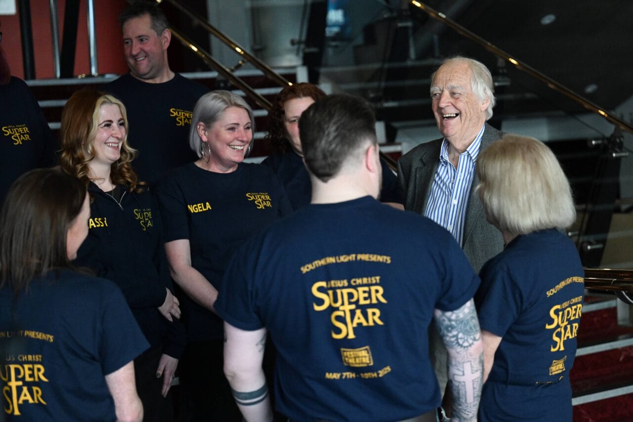 A group of people wearing "Jesus Christ Superstar" shirts stand together and smile, talking with an older man in a suit inside a building.