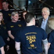A group of people wearing "Jesus Christ Superstar" shirts stand together and smile, talking with an older man in a suit inside a building.