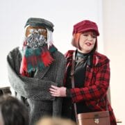A woman in a red plaid coat and hat stands next to a life-sized figure dressed in a coat, scarf, cap, and glasses, with a yarn beard and mustache.
