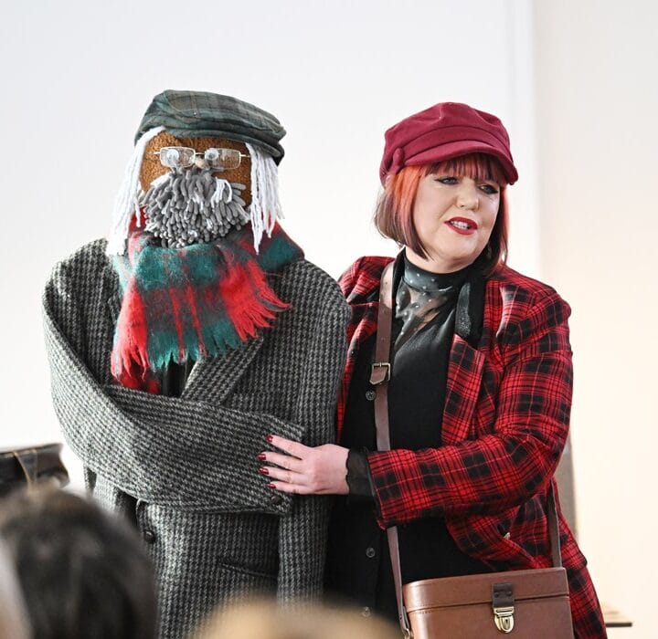 A woman in a red plaid coat and hat stands next to a life-sized figure dressed in a coat, scarf, cap, and glasses, with a yarn beard and mustache.