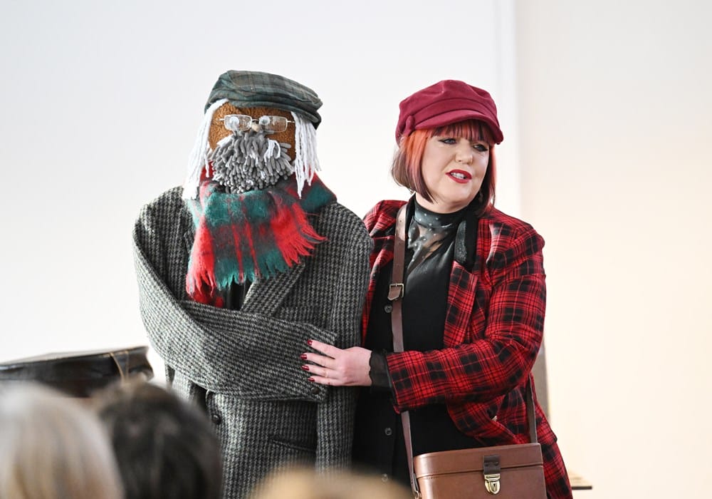 A woman in a red plaid coat and hat stands next to a life-sized figure dressed in a coat, scarf, cap, and glasses, with a yarn beard and mustache.