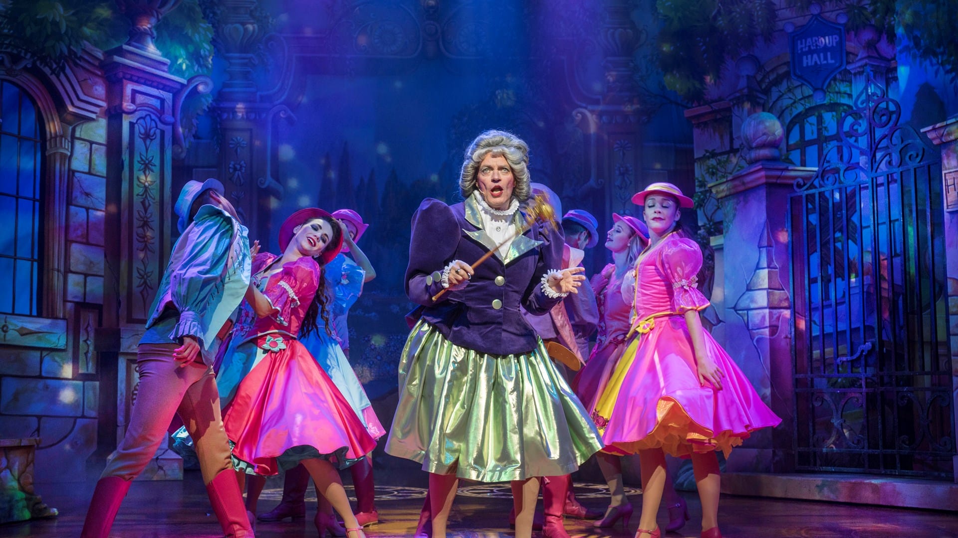 A group of performers in colorful costumes sing and dance on a brightly lit stage set resembling a whimsical garden scene.
