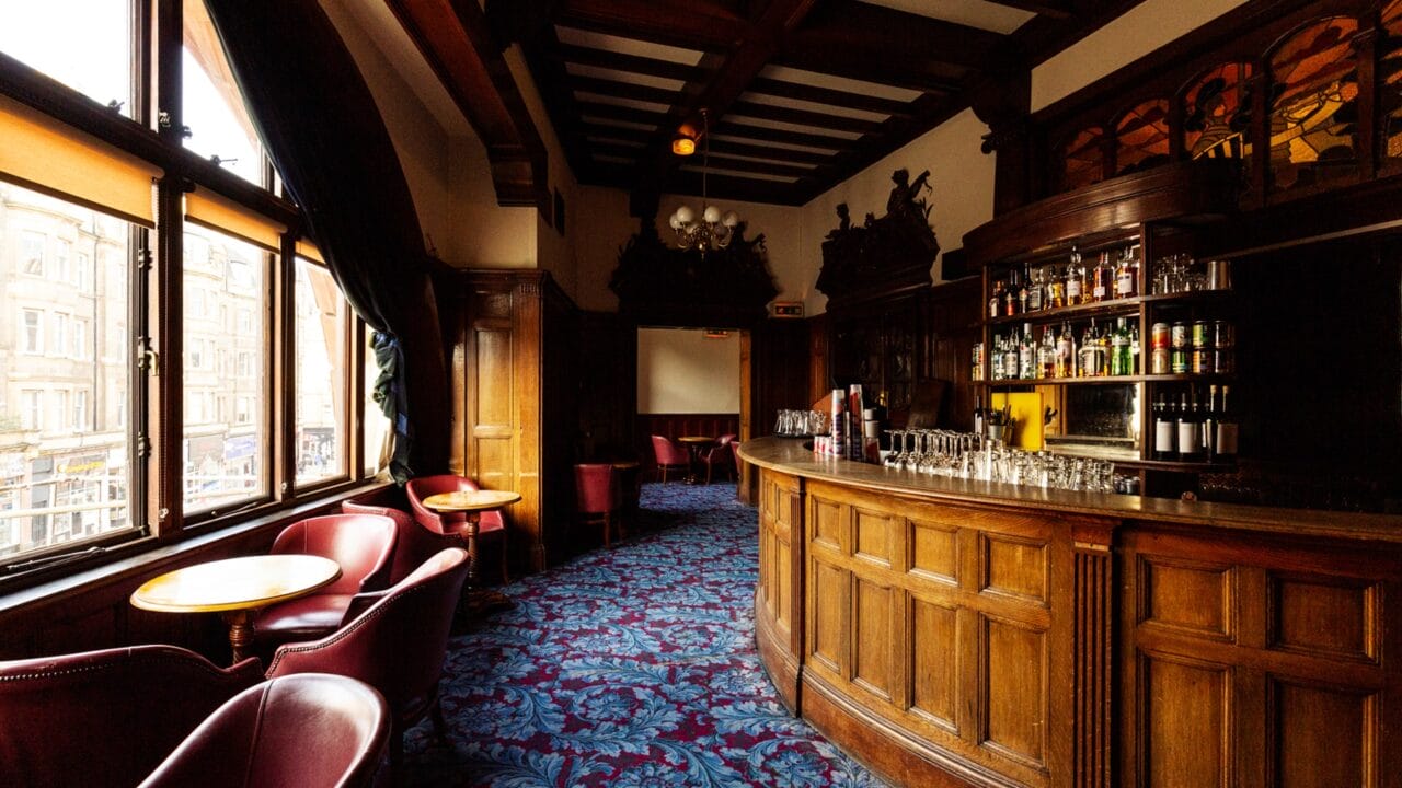 A traditional pub interior with wooden bar, shelves of bottles, blue patterned carpet, round tables, and chairs by large windows overlooking a city street.