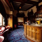 A traditional pub interior with wooden bar, shelves of bottles, blue patterned carpet, round tables, and chairs by large windows overlooking a city street.