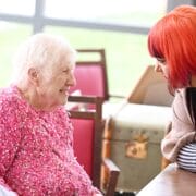 A woman with red hair leans on a table, talking to two elderly women sitting next to each other, one wearing a pink sweater, indoors.