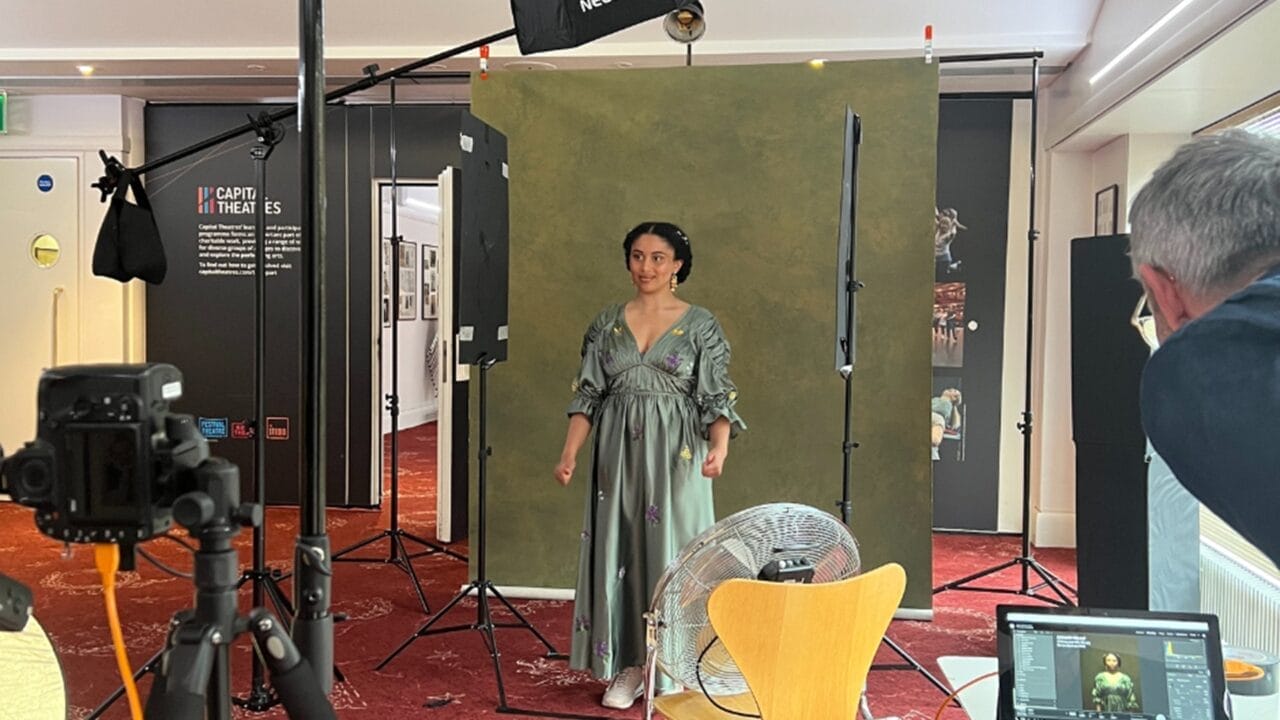 Danielle Jam poses in a green dress under the spotlight, framed by a green backdrop in a photo studio setup, with lights, camera, fan, and a photographer adjusting equipment.