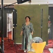 Danielle Jam poses in a green dress under the spotlight, framed by a green backdrop in a photo studio setup, with lights, camera, fan, and a photographer adjusting equipment.