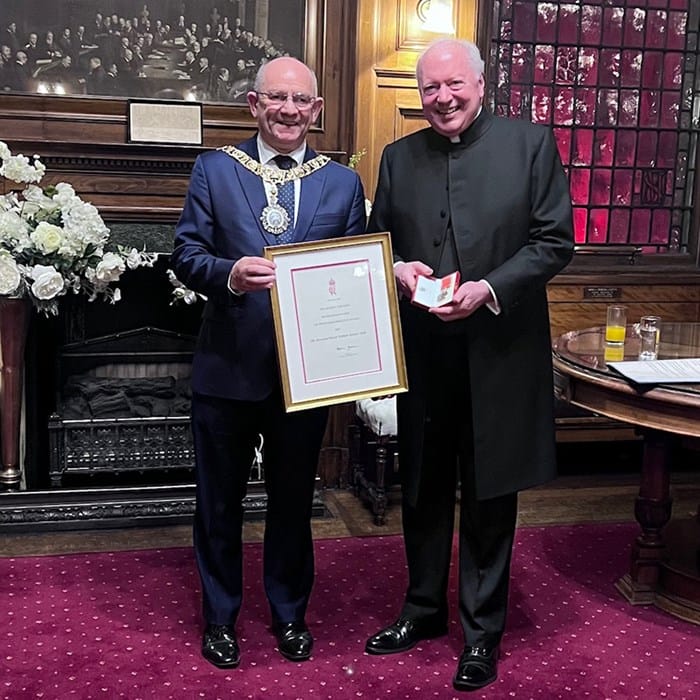 Two men in formal attire stand indoors; one holds a framed certificate, and the other holds a box. They are smiling and posing for a photo in an ornate room.