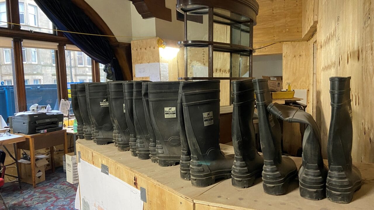 A row of black rubber boots is lined up on a wooden counter in a room under construction or renovation, with office supplies and blueprints visible in the background.