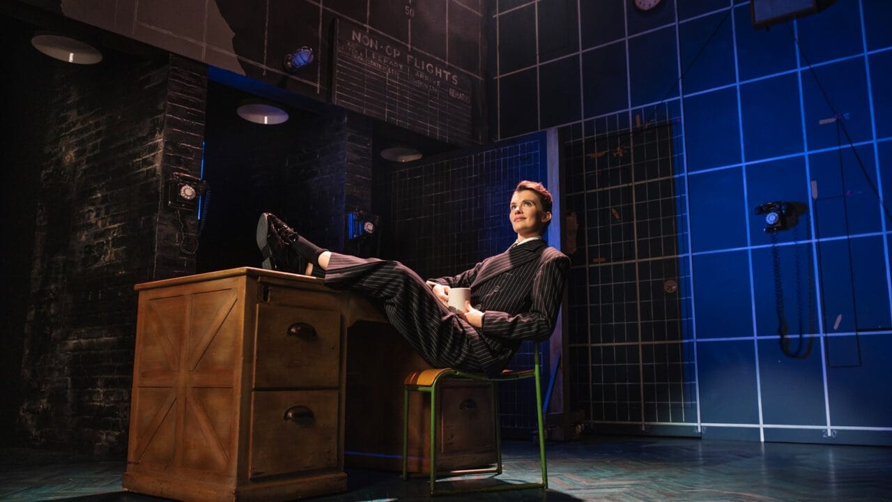 A person in a pinstripe suit sits at a desk with their feet up, holding a mug, on a stage with a blue and black background and visible telephones on the walls.