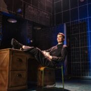 A person in a pinstripe suit sits at a desk with their feet up, holding a mug, on a stage with a blue and black background and visible telephones on the walls.