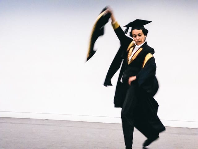 A person in graduation attire dances energetically, holding their cap and gown against a plain, light-colored background.