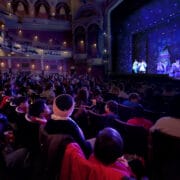 Audience members watch a theatrical performance on stage inside a large, ornate theatre with balconies and decorative lighting.