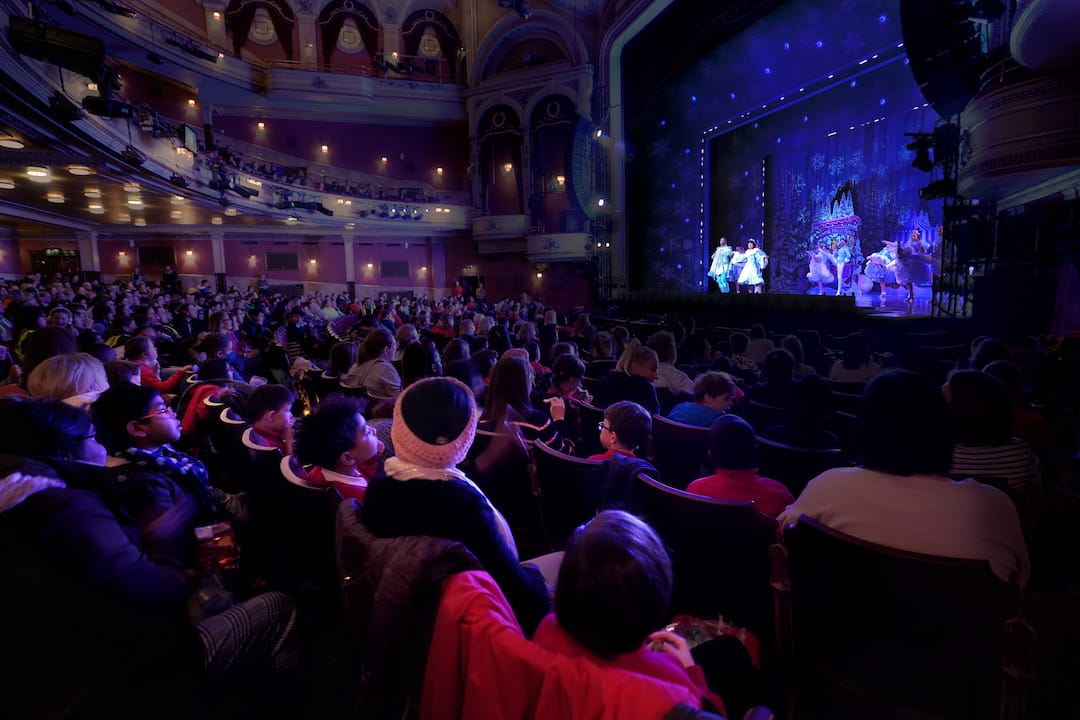 Audience members watch a theatrical performance on stage inside a large, ornate theatre with balconies and decorative lighting.