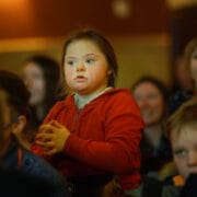 A young girl in a red hoodie sits among a group of people, appearing attentive and focused on something off-camera.