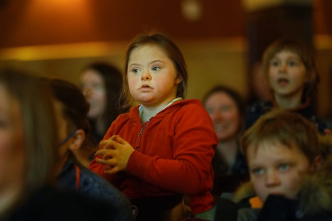 A young girl in a red hoodie sits among a group of people, appearing attentive and focused on something off-camera.