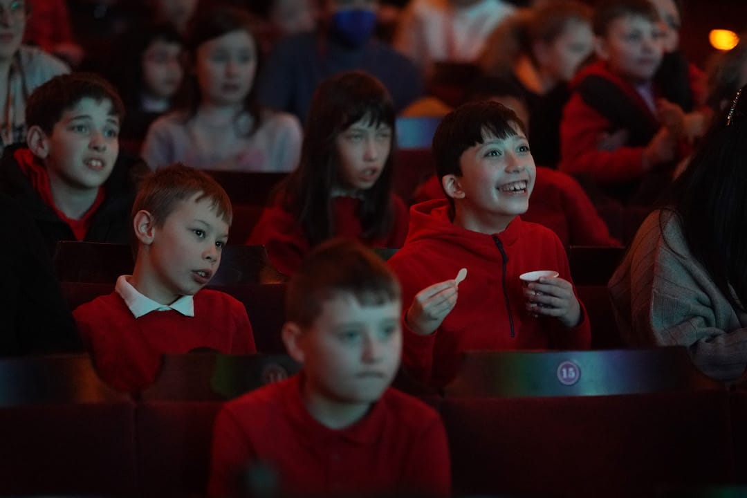 A group of children in red clothing sit in theatre seats, watching something on stage with various facial expressions of interest and excitement.