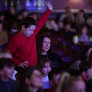 A boy in a red shirt stands and raises his hand in a seated audience, while people around him watch attentively.