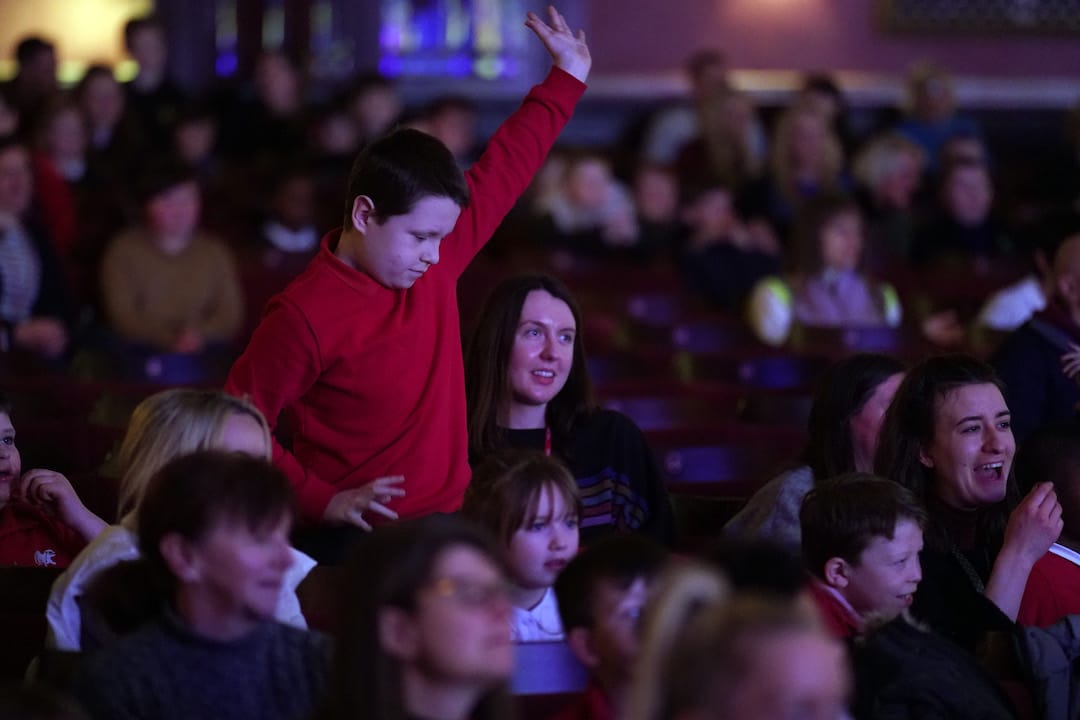 A boy in a red shirt stands and raises his hand in a seated audience, while people around him watch attentively.