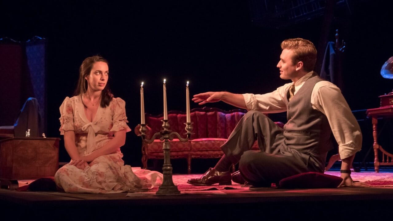 Two actors sit on the floor near a lit candelabrum on stage, dressed in vintage-style clothing, with antique furniture and dim lighting in the background.