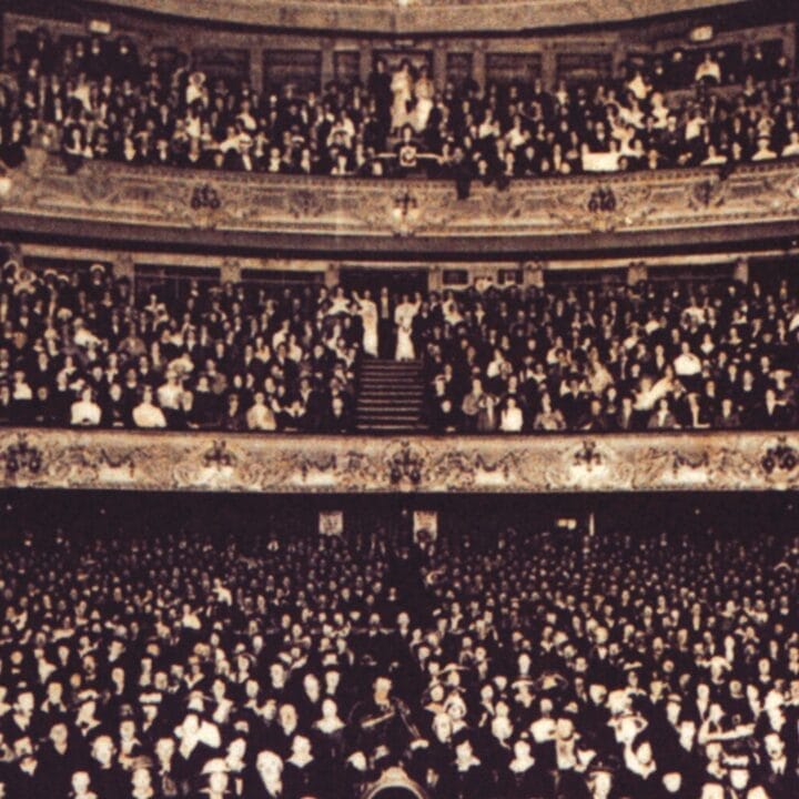 A large audience fills the seats and balconies of the ornate King’s Theatre, facing the stage in a sepia-toned photo.