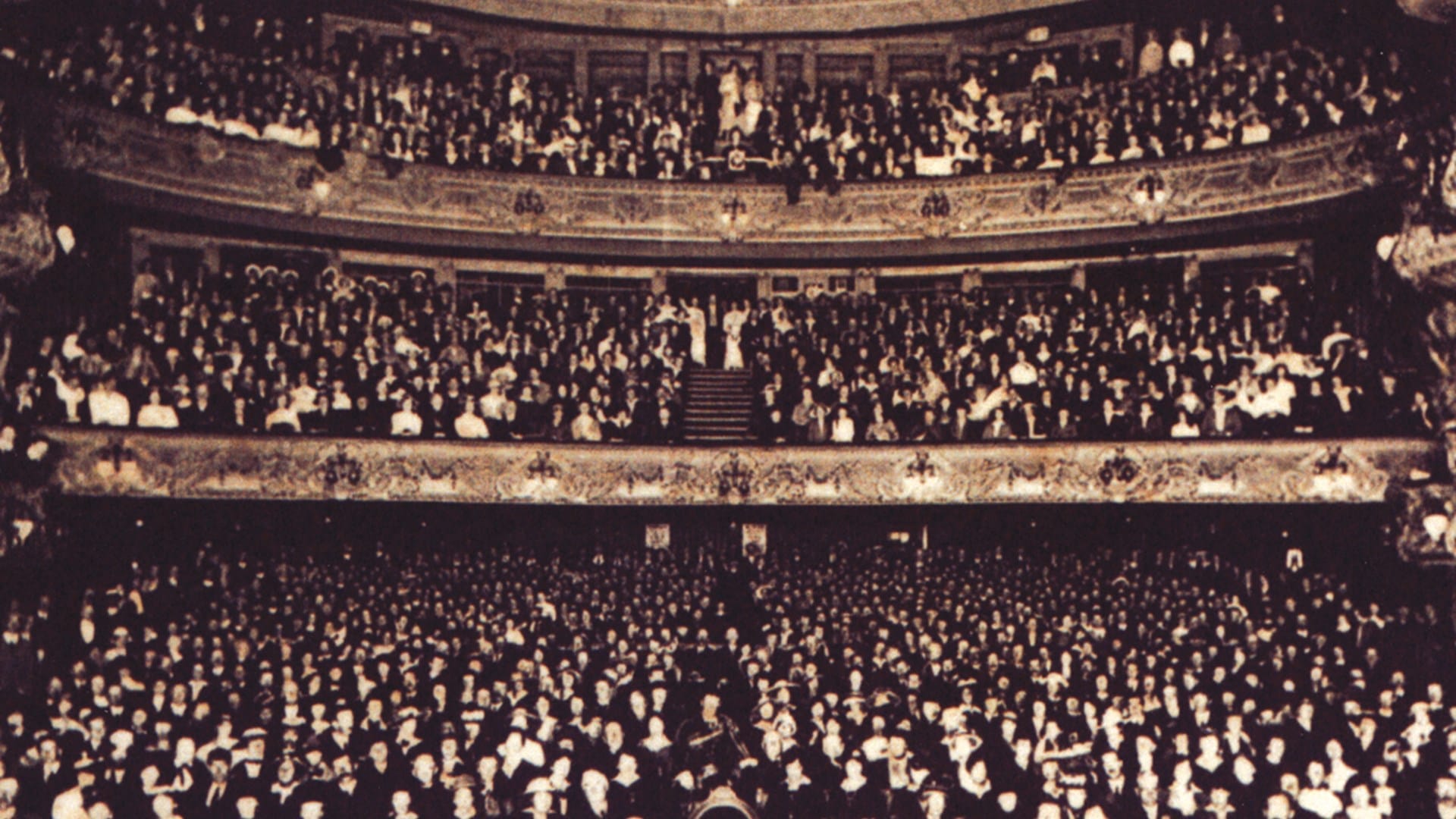 A large audience fills the seats and balconies of the ornate King’s Theatre, facing the stage in a sepia-toned photo.