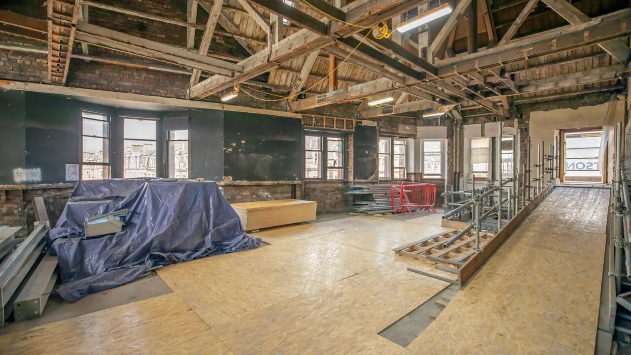 A large room under renovation with exposed wooden beams, construction materials, plywood flooring, and windows lining the far wall.