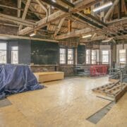A large room under renovation with exposed wooden beams, construction materials, plywood flooring, and windows lining the far wall.