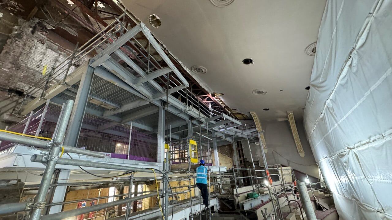 A construction worker stands on scaffolding inside a large building under renovation, with exposed pipes and partially finished walls visible.